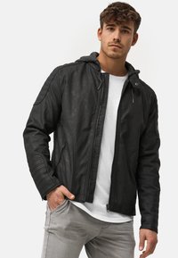 Schwarze Lederjacke mit grauem Kapuzenaufsatz, ausgestattet mit gerippten Schulterakzenten und zwei Reißverschlusstaschen, kombiniert mit einem weißen T-Shirt und hellgrauen Hosen.