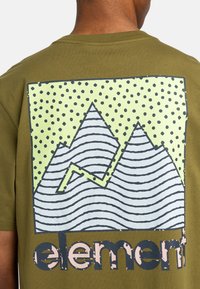 Olivegrünes T-Shirt mit einem großen Grafikdesign von Bergen in hellgrün und marineblau, verziert mit gepunkteten Akzenten und dem Wort "Element" darunter.