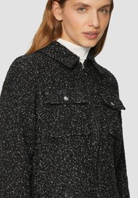 Schwarze und weiße strukturierte Jacke mit einem Reißverschluss vorne, Kragen und zwei Brusttaschen. Besitzt ein gesprenkeltes Muster und weichen Stoff.