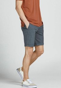 Pantaloni shorts da uomo grigio scuro con una vestibilità elegante, caratterizzati da tasche frontali, orlo piegato e una texture sottile. Indossati con una maglietta color ruggine e scarpe chiare.