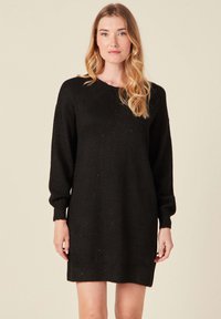 Svart stickad klänning med rund halsringning, långa ärmar och ribbade muddarna. Mjuk textur och en lös passform, med subtila metallisk sprickor.