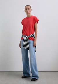 Woman wearing a red t-shirt, light blue wide-leg jeans, black shoes, and a black-and-white striped sweater tied around her waist.