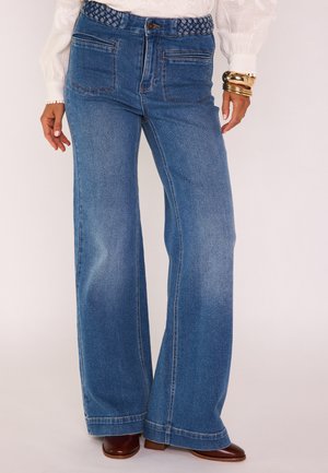 Femme portant un jean bleu à jambes larges avec poches avant, une ceinture tressée, un chemisier blanc, des bracelets dorés et des chaussures à talons marron.