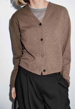 Cardigan en laine marron avec un col en V, comportant cinq boutons noirs, des poignets côtelés et un design court. Porté par-dessus une chemise grise.
