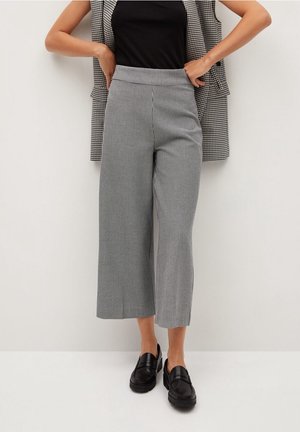 Kobieta w czarnym topie, czarno-białych kratkowanych krótkich spodniach z szerokimi nogawkami, czarnych mokasynach i kratkowanej marynarce zarzuconej na ramiona.