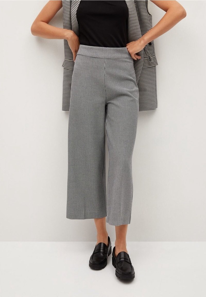 Femme portant un haut noir, un pantalon large court à carreaux noir et blanc, des mocassins noirs et un blazer à carreaux posé sur les épaules.