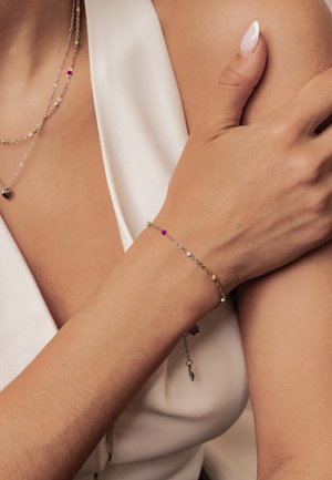 Feines Goldketten-Armband mit kleinen bunten Perlen, getragen am Handgelenk, Hand ruht auf der gegenüberliegenden Schulter, weißes ärmelloses Top sichtbar.