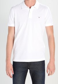 Polo shirt blanc à manches courtes en coton, avec un col, deux boutons et un petit logo sur la poitrine gauche. Accompagné d'un jean sombre.