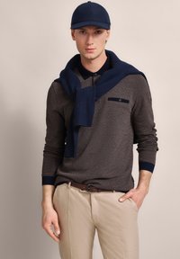 Dunkelbraunes Langarm-Polohemd mit marineblauem Rundhalsausschnitt und gerippten Bündchen, mit einer Brusttasche und einem marineblauen Pullover, der über die Schultern drapiert ist. Helle Hose.