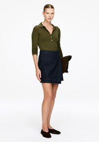 Woman wearing an olive green ribbed long-sleeve top, dark blue denim wrap skirt, black flats, holding a brown clutch bag.