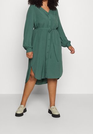 Robe chemise verte à boutons avec un col en V, taille cintrée, manches longues bouffantes, fentes latérales et texture lisse. Portée avec des mocassins de couleur claire.