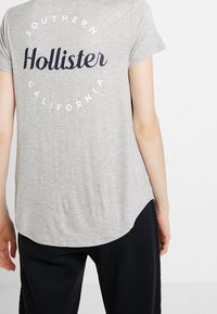 Šedé tričko s krátkým rukávem a zaobleným lemem, na zadní straně s námořnickou kulatou grafikou s nápisem "Hollister Southern California".