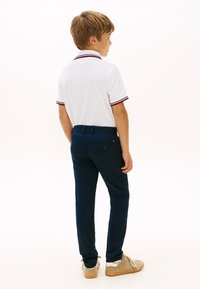 Ragazzo in piedi di profilo, indossa una polo bianca, pantaloni blu scuro e sneakers beige su uno sfondo bianco uniforme.