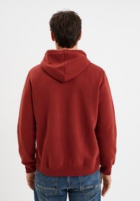 Sudadera con capucha roja, de mangas largas y puños acanalados, que muestra una tela suave y texturizada. La parte posterior presenta un diseño de bolsillo tipo canguro.