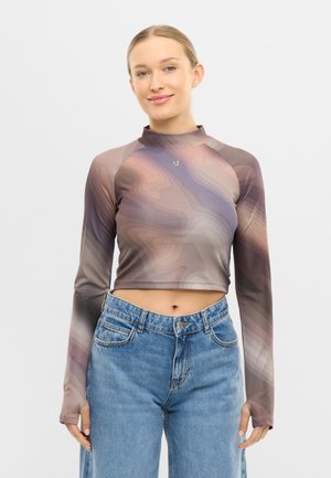 Mosolygó nő szőke hajjal, hosszú ujjú, kivágott felsőt visel, amelyen absztrakt lila és bézs minták találhatók, valamint kék, magas derekú farmert.