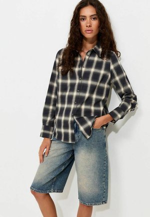 Femme aux longs cheveux bouclés portant une chemise à carreaux noire et blanche et un short en jean bleu délavé jusqu’au genou, une main dans la poche.