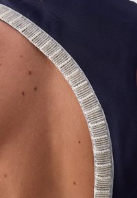 Gros plan d'une épaule nue à côté d'un tissu foncé orné de perles blanches et de fils argentés. Plusieurs petits grains de beauté sont visibles sur la peau.