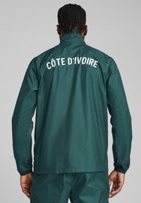Green, lightweight jacket with high collar and long sleeves. Features "CÔTE D'IVOIRE" printed in white on the back. Smooth texture.
