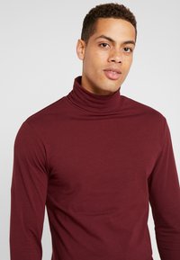 Tricou cu maneci lungi de culoare burgundy, dintr-un material neted, cu un design ajustat și un guler înalt pentru a oferi căldură la nivelul gâtului.