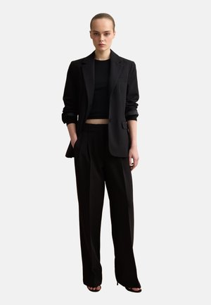 Woman with hair tied back wearing a black blazer with rolled sleeves, black crop top, wide-leg black trousers, and black open-toe heels.