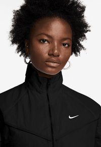 Casaco preto com fecho zip, colarinho alto, apresentando o logo branco da Nike, feito de um tecido suave. Mangas curtas e design ajustado.