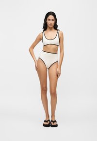 Femme debout sur fond blanc portant un bikini blanc taille haute avec une bordure noire et des sandales plateformes noires.