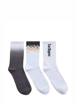 Trzy pary skarpetek: jedna z ciemnoszarym gradientowym wzorem, jedna biała z beżowym wzorem kropli, a jedna gładka biała z czarnym napisem.