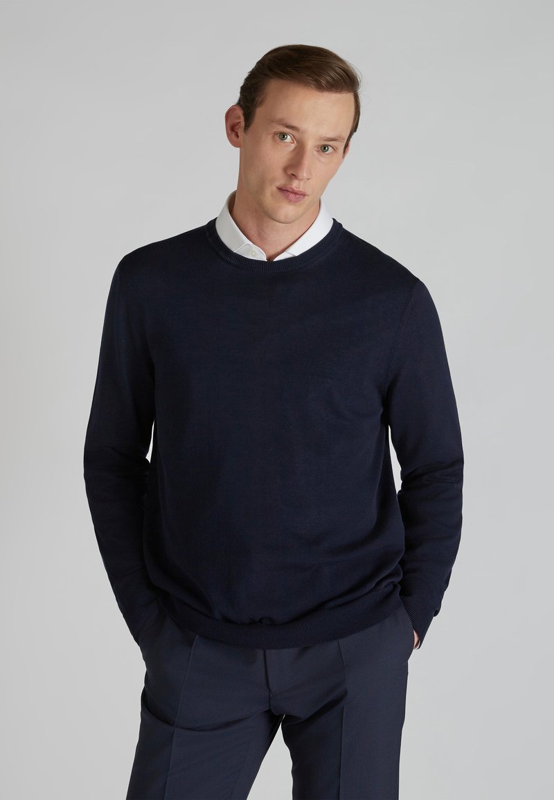 Navy blue knitted sweater with a round neck, ribbed cuffs, and hem. Worn over a white collared shirt, smooth texture, classic fit.