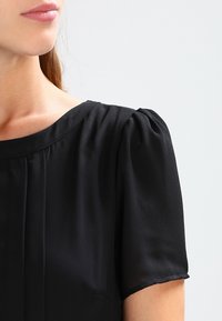 Černá blůza s krátkým rukávem a kulatým výstřihem, vyrobená z jemné, splývavé látky a s jemnými záhyby vpředu. Hladká textura.