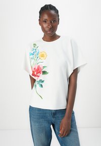 White floral top featuring a vibrant pink, yellow, and blue flower design, short sleeves, and a relaxed fit, paired with denim trousers.