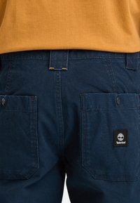 Navy blue cargo shorts made of sturdy fabric, featuring two back pockets and a black Timberland logo patch. Orange stitching accents noticeable.