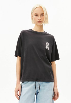 Junge Person mit kurzen blonden Haaren, die ein lockeres schwarzes T-Shirt mit einem weißen Schlüsselloch-Symbol auf der Brust und hellblaue Jeans trägt.