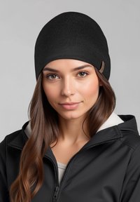Zwarte gebreide muts met een gladde textuur, aansluitend ontwerp en een klein leren logolabel; gedragen bij een jack met ritssluiting.