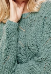 Eine blonde Person trägt einen strukturierten, hellgrünen Strickpullover und berührt mit einer Hand ihren Hals.