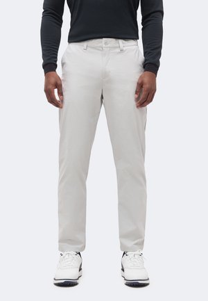 Homme portant un pantalon gris clair, un t-shirt noir à manches longues et des baskets blanches, debout devant un fond uni clair.