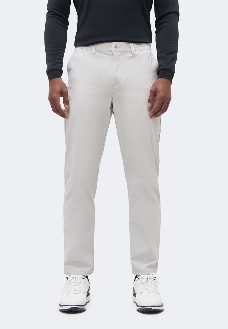 Homme portant un pantalon gris clair, un t-shirt noir à manches longues et des baskets blanches, debout devant un fond uni clair.