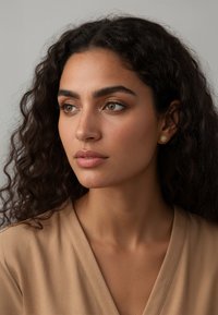 Goldene, blumige Steckerohrringe mit strukturierter Oberfläche, getragen von einer Person mit lockigem Haar, neutral gefärbtem Oberteil, neutralem Hintergrund.