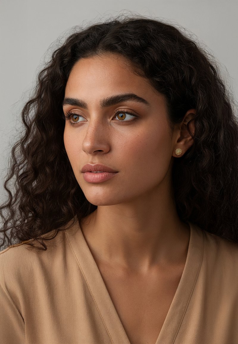 Goldene, blumige Steckerohrringe mit strukturierter Oberfläche, getragen von einer Person mit lockigem Haar, neutral gefärbtem Oberteil, neutralem Hintergrund.