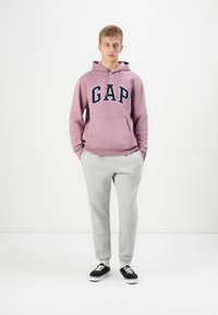 Purple hooded sweatshirt with "GAP" logo in navy, paired with light gray sweatpants and black sneakers with white soles. Simple design.