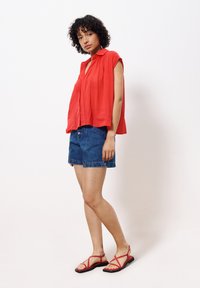 Rote, ärmellose Bluse aus leichtem Stoff; kombiniert mit dunklen Denim-Shorts mit zwei vorderen Knöpfen und flachen, roten Sandalen mit Riemen.