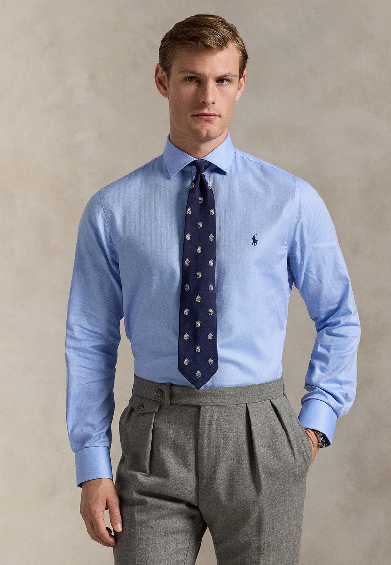 Cămașă în dungi bleu deschis cu guler ascuțit, combinată cu o cravată bleumarin închis cu accente aurii și pantaloni gri plisati.