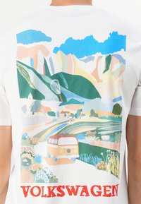 Hvid bomulds t-shirt med et farverigt landskabsgraphics med bjerge og en rød Volkswagen varevogn. "VOLKSWAGEN" trykt med fede røde bogstaver.
