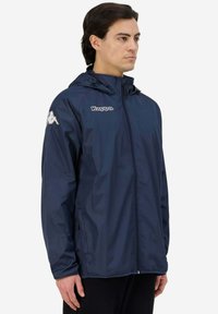 Giacca Kappa blu navy con zip frontale, cappuccio regolabile e dettagli con logo. Materiale leggero con una texture liscia e maniche lunghe.