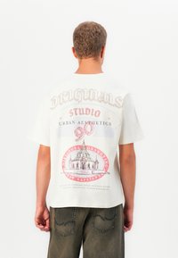 Asmuo vilki baltą marškinėlį su spalvinga grafika ir tekstu, įskaitant "Originals," "Studio," "Urban Aesthetics," ir pastato iliustraciją.