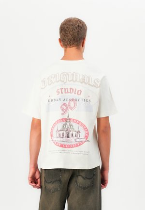 Person iført en hvid T-shirt med farverig grafik og tekst, herunder "Originals," "Studio," "Urban Aesthetics," og en bygning illustration.