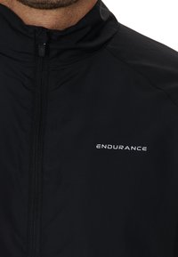 Svart jacka med hög krage, med en framdragkedja och ordet "ENDURANCE" i vitt på vänster sida. Slät, lättviktsmaterial.