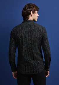 Černá košile s modrým svislým vzorem textu, dlouhými rukávy a límečkem na knoflíky, zobrazená zezadu na modrém pozadí.