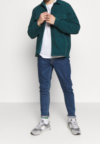 Teal cordflätad skjortjacka med två bröstfickor, tillsammans med åtsittande blå jeans och grå sneakers med vita detaljer.