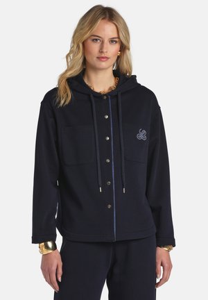 Femme portant un sweat à capuche noir avec boutons-pression sur le devant, deux poches poitrine, logo brodé et bijoux en or sur fond blanc.