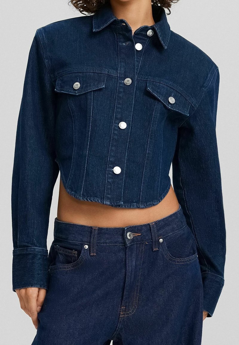 Mörkblå denimjacka i cropped model med två bröstfickor, silverfärgade tryckknappar och klassisk krage; matchad med mörka jeans.